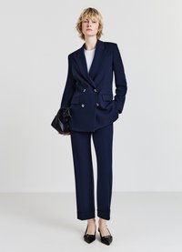 Blazer doppiopetto blu navy con bottoni dorati, abbinato a pantaloni sartoriali coordinati e ballerine nere a punta. Gli accessori includono una borsa nera.