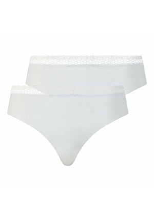 Femme debout, de face, portant un crop top noir sans manches et un sous-vêtement taille haute blanc avec une bordure en dentelle, sur un fond uni.