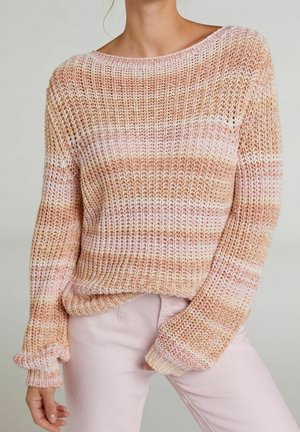 Femme portant un pull ample à mailles lâches avec des rayures horizontales roses, beige et blanches, associé à un pantalon rose clair sur un fond neutre.