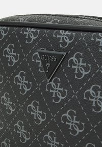 Geantă sintetică neagră cu un model matlasat diagonal, decorată cu logo-uri argintii, finisată cu un emblema triunghiulară Guess din metal.
