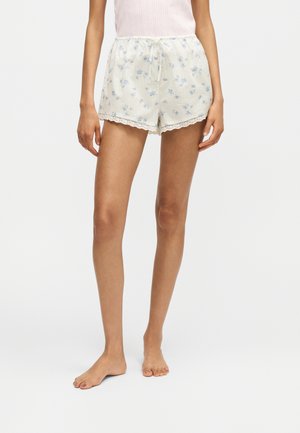 Vrouw draagt witte korte broek met blauwe bloemenprint en kanten rand, gecombineerd met een lichtroze mouwloos topje, staat op blote voeten.