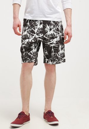 Mężczyzna w czarnych szortach z białym abstrakcyjnym wzorem rozprysków, białej koszuli z długim rękawem i czerwonych trampkach stoi na jednolitym tle.