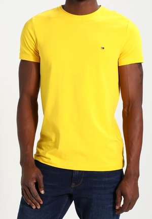 Mężczyzna ubrany w prostą żółtą koszulkę na krótki rękaw z małym logo Tommy Hilfiger na piersi, w połączeniu z ciemnoniebieskimi dżinsami.