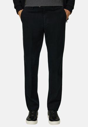 Mand iført sorte bukser med lige pasform med hænderne i lommerne og sorte sneakers med hvide såler mod en ensfarvet baggrund.