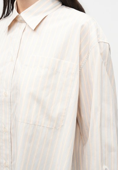Γυναίκα με λευκό πουκάμισο με λεπτές κάθετες μπεζ ρίγες και τσέπη στο στήθος, εμφανιζόμενη από τον ώμο έως το μέσο του κορμού.