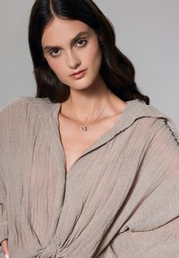 Srebrny naszyjnik o splątanym wzorze noszony na jasnobeżowej, teksturowanej bluzce z luźnym, drapowanym krojem i złożonym kołnierzem.