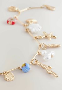 Guldtonet armbånd med forskellige charms, herunder muslingeskaller, søstjerner og farverige perler, i en blanding af glansfulde og teksturerede overflader.
