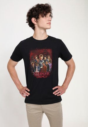 Zwart katoenen T-shirt met een grafische afbeelding van personages uit "Stranger Things" voor een rode, gestructureerde achtergrond. Korte mouwen.