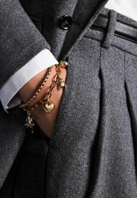 Grå uldblazer med enkelt knap, hvid skjortearm der stikker ud, prydet med lag på lag guldcharmsarmbånd inklusive hjerter og frøer.