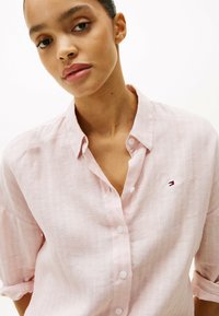Camisa de botón rosa claro hecha de una tela texturizada, con un pequeño logo bordado en el pecho y mangas enrolladas.