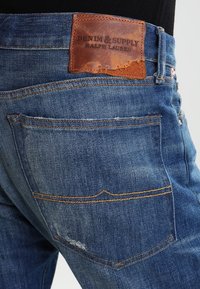 Jeans de mezclilla azul oscuro con un parche de cuero que dice "DENIM & SUPPLY RALPH LAUREN". Presenta cinco bolsillos y detalles de costura en contraste.