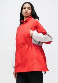 Jachetă roșie și albă cu glugă, talie elastică, mâneci lungi și buzunar frontal. Materialul pare ușor și fin.