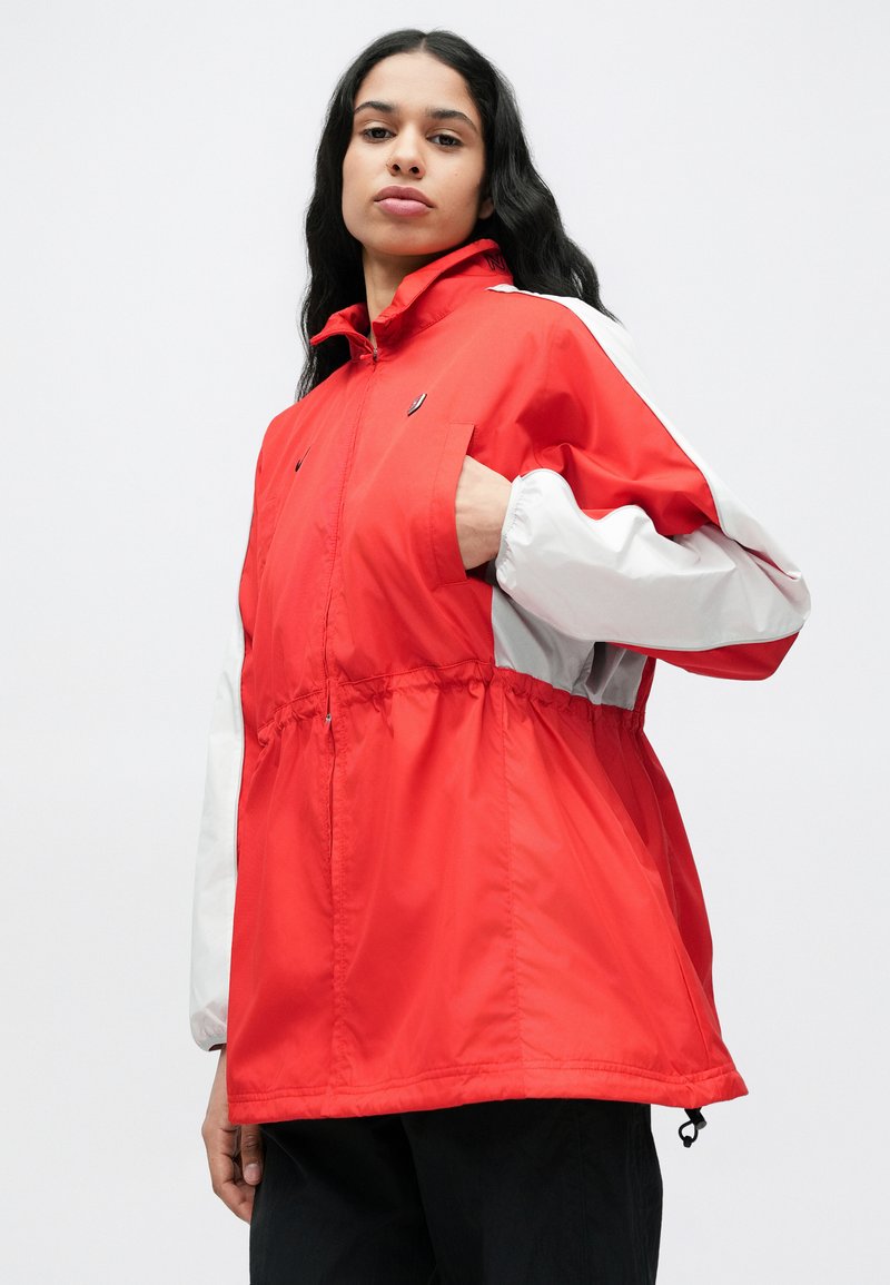 Rød og hvid hættejakke med elastik i taljen, lange ærmer og lynlåslomme foran. Stoffet ser let og glat ud.