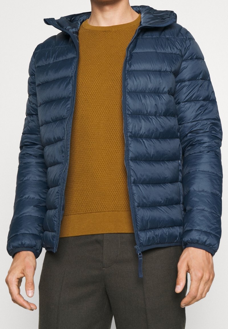 Doudoune marine avec capuche, présentant un design matelassé, une fermeture éclair à l'avant et des poignets élastiques. Portée par-dessus un pull texturé moutarde.