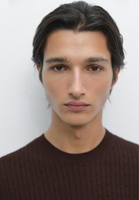 Portrét mladého muže s tmavými, vlnitými vlasy. Má na sobě texturovaný tmavě hnědý svetr a za ním je neutrální pozadí. Hladká pleť a hnědé oči.