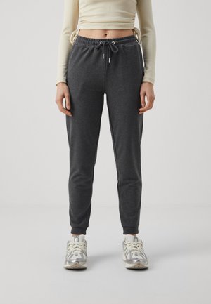 Mujer con pantalones jogger gris oscuro y zapatillas blancas, de pie frente a un fondo liso gris claro.