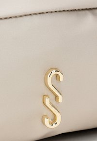 Béžová taška s hladkým, štruktúrovaným povrchom a zlatým kovovým doplnkom v tvare stylizovaného písmena "S". Viditeľné embosované logo.
