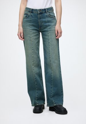 Personne portant un jean bleu taille haute à jambes larges avec un effet délavé, un haut blanc rentré, et des chaussures plateformes noires épaisses.