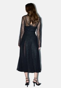 Abito lungo in pizzo nero con maniche lunghe, corpetto aderente, schiena scoperta e gonna morbida con dettagli scintillanti e spacchi laterali.