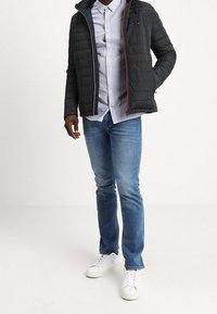 Svart vadderad jacka med huva, dragkedjestängning, röda detaljer, ljusblå skjorta med knappar, blå jeans och vita sneakers.
