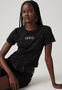 Schwarz Baumwoll-T-Shirt mit kurzen Ärmeln, ausgestattet mit weißem "LEV'S"-Logo auf der Brust. Getragen mit schwarzen Jeans-Shorts in Sitzposition.