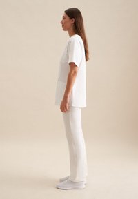 Top bianco in lino con maniche corte e due tasche, abbinato a leggings bianchi e sneakers chiare. Texture liscia, vestibilità rilassata, design minimalista.
