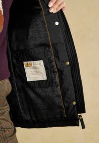 Jacheta neagră matlasată are la interior un căptușeală cu un model, un buzunar cu etichetă și accente aurii. Detalii cu fermoar și închidere cu capse sunt vizibile.