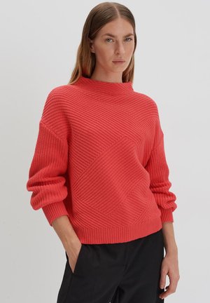 Coral knit sweater with a high neck, loose fit, and textured diamond patterns. Features oversized sleeves and ribbed cuffs.