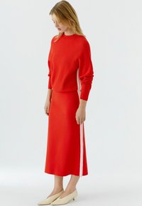 Pull en tricot rouge associé à une jupe longue assortie avec des bandes blanches sur les côtés. Le modèle porte des chaussures beige à petit talon.