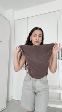 Ung kvinde i grå shorts holder en brun ærmeløs top strakt mellem sine hænder i et lyst, minimalistisk rum.