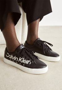 Černé kožené tenisky s bílou gumovou podrážkou, s výrazným logem "Calvin Klein Jeans" na boku a klasickým šněrováním.