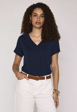Donna con capelli ricci che indossa una maglietta blu navy con scollo a V e maniche corte, pantaloni bianchi, cintura marrone e braccialetti d'oro su sfondo semplice.