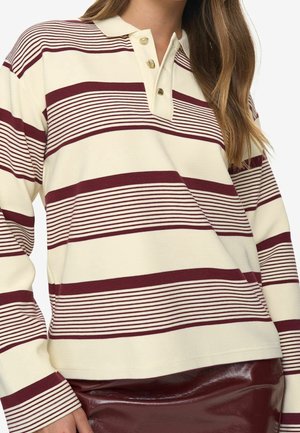 Femme portant une chemise à manches longues rayée crème et bordeaux avec un col et trois boutons, assortie à une jupe en cuir bordeaux.