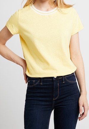 T-shirt gialla a maniche corte con colletto girocollo bianco, realizzata in un materiale morbido, abbinata a jeans in denim blu scuro.