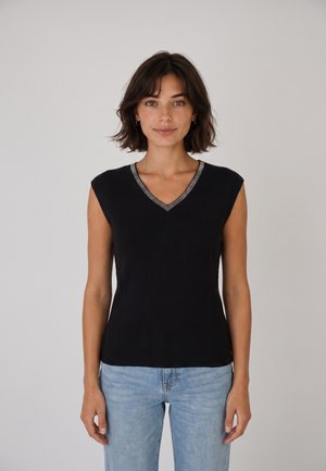 Vrouw met kort donker haar, gekleed in een mouwloos zwart topje met een zilverkleurige V-hals en lichtblauwe spijkerbroek, staand tegen een effen achtergrond.