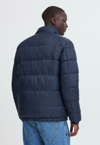 Marineblå puffjakke med ståkrave, quiltet design og elastiske manchetter. Glat tekstur og let skinnende finish.
