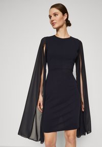 Ναυτικό φόρεμα με εφαρμοστή σιλουέτα και διάφανα, ρέουσα μανίκια τύπου κάπας. Λείο ύφασμα; μέχρι το γόνατο με στρογγυλή λαιμόκοψη.