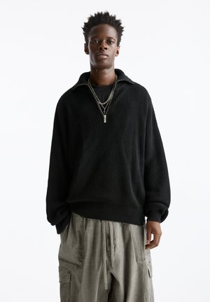 Black knitted sweater with a high collar and zip detail, paired with light grey cargo pants featuring multiple pockets and drawstring.