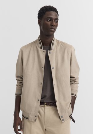 Mann trägt beige perforierte Bomberjacke über dunklem Hemd und beigefarbenen Hosen, steht mit einer Hand in der Tasche vor einfarbigem Hintergrund.