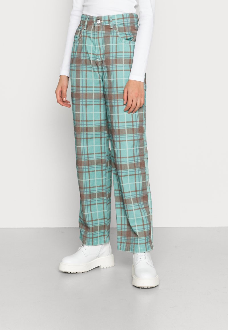 Calças xadrez em verde-azulado e castanho, com um ajuste descontraído, bolsos e um tecido ligeiramente texturizado. Combinadas com sapatos de plataforma brancos.