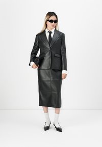 Kvinne iført et svart lærblazer og skjørt, hvit skjorte og sokker, svart slips og solbriller, som holder en svart kuvertveske.