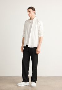 Hvit skjorte over en hvit T-skjorte, kombinert med svarte bukser og hvite sneakers. Glatt stoff, rene linjer og minimalistisk design.