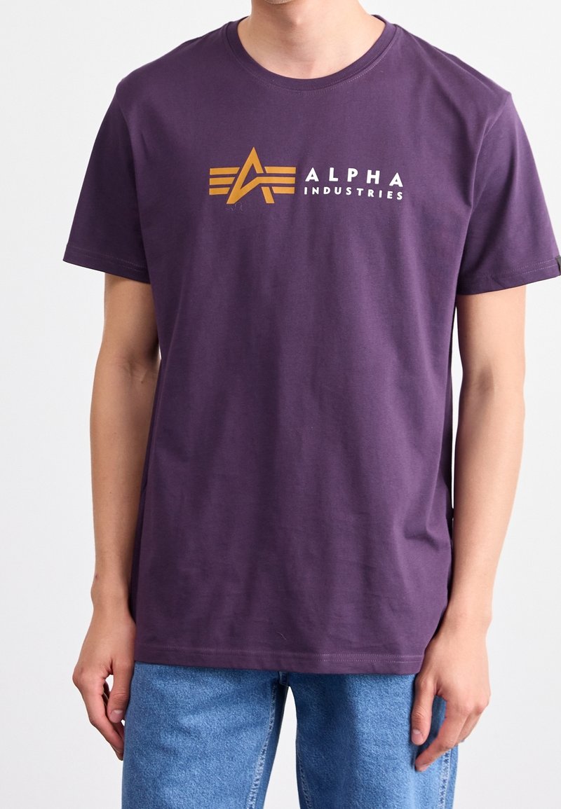 Fialové bavlněné tričko s krátkými rukávy. Obsahuje logo v oranžové a bílé barvě, zobrazující abstraktní design hvězdy, umístěný na středu hrudníku.