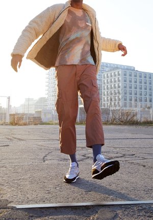 Beige schapenvacht jacket over een tie-dye t-shirt, roestkleurige cargobroek met zakken, en zwart-witte sneakers op een asfalt achtergrond.