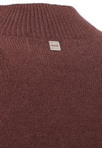 Maglione marrone a maglia con colletto a costine e una piccola targhetta metallica con testo in rilievo vicino al collo sul retro.