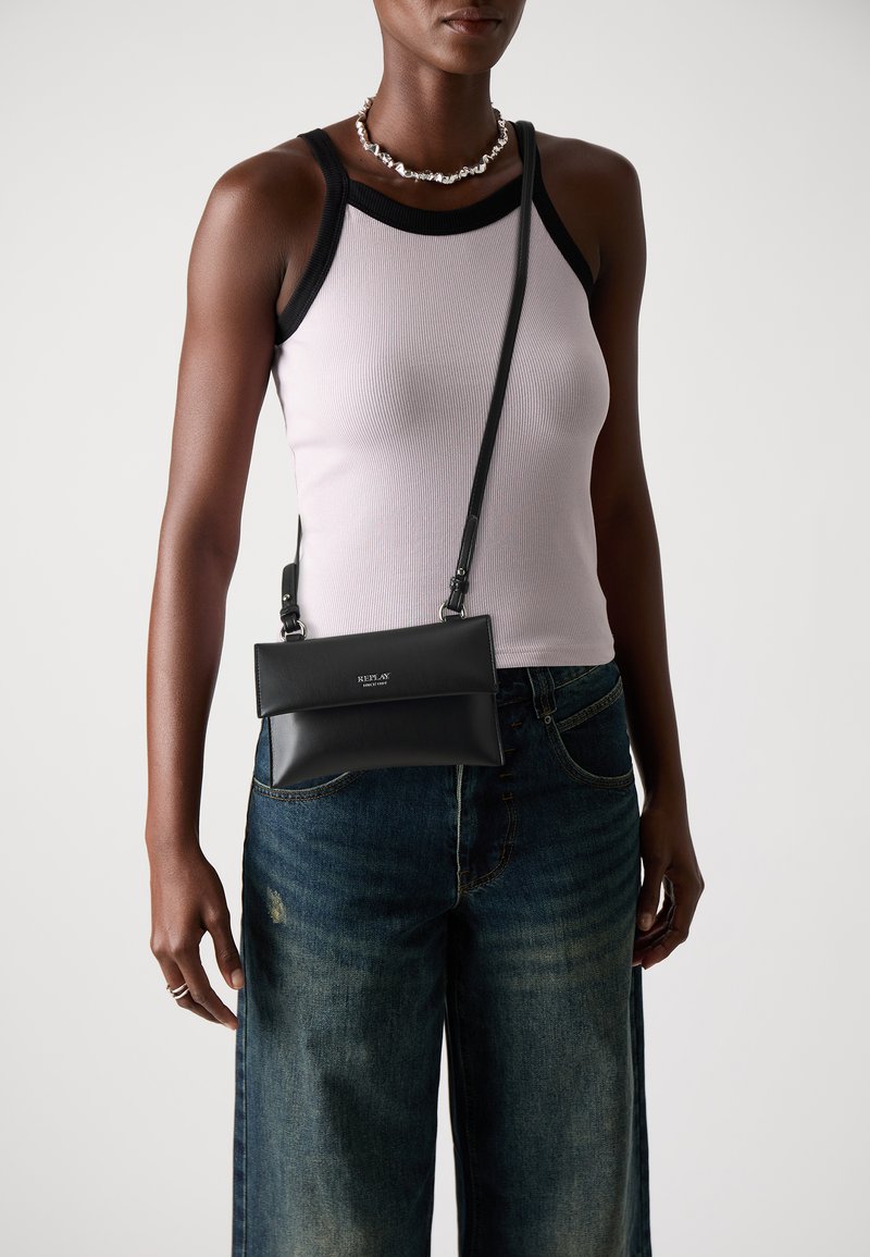 Czarna skórzana torebka crossbody o minimalistycznym designie, z klapką i srebrnymi akcentami, noszona na różowej, prążkowanej koszulce na ramiączkach oraz dżinsowych spodniach.