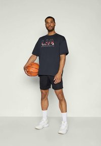 Sort kortærmet T-shirt med "UA BASKETBALL" grafik, parret med sorte shorts. Modellen holder en basketball og har hvide sneakers på.