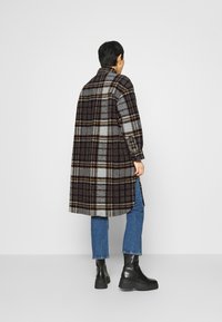 Lunga giacca a quadri in tonalità di grigio, nero e giallo. Realizzata in tessuto di lana con una vestibilità ampia e fessure laterali. Indossata sopra jeans blu e stivali neri.