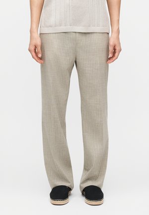 Persona che indossa pantaloni larghi color beige chiaro a righe sottili e espadrillas nere slip-on con suole intrecciate naturali, in piedi contro uno sfondo bianco.