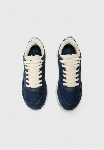 Ténis azul-escuro com atacadores e sola crèmes, apresentando um design texturizado e um detalhe em verde no logo na lateral. Materiais suaves e acolchoados foram utilizados.